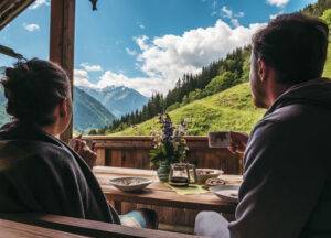 Ein Paar sitzt vor einer Berghütte, frühstückt und genießt die Landschaft