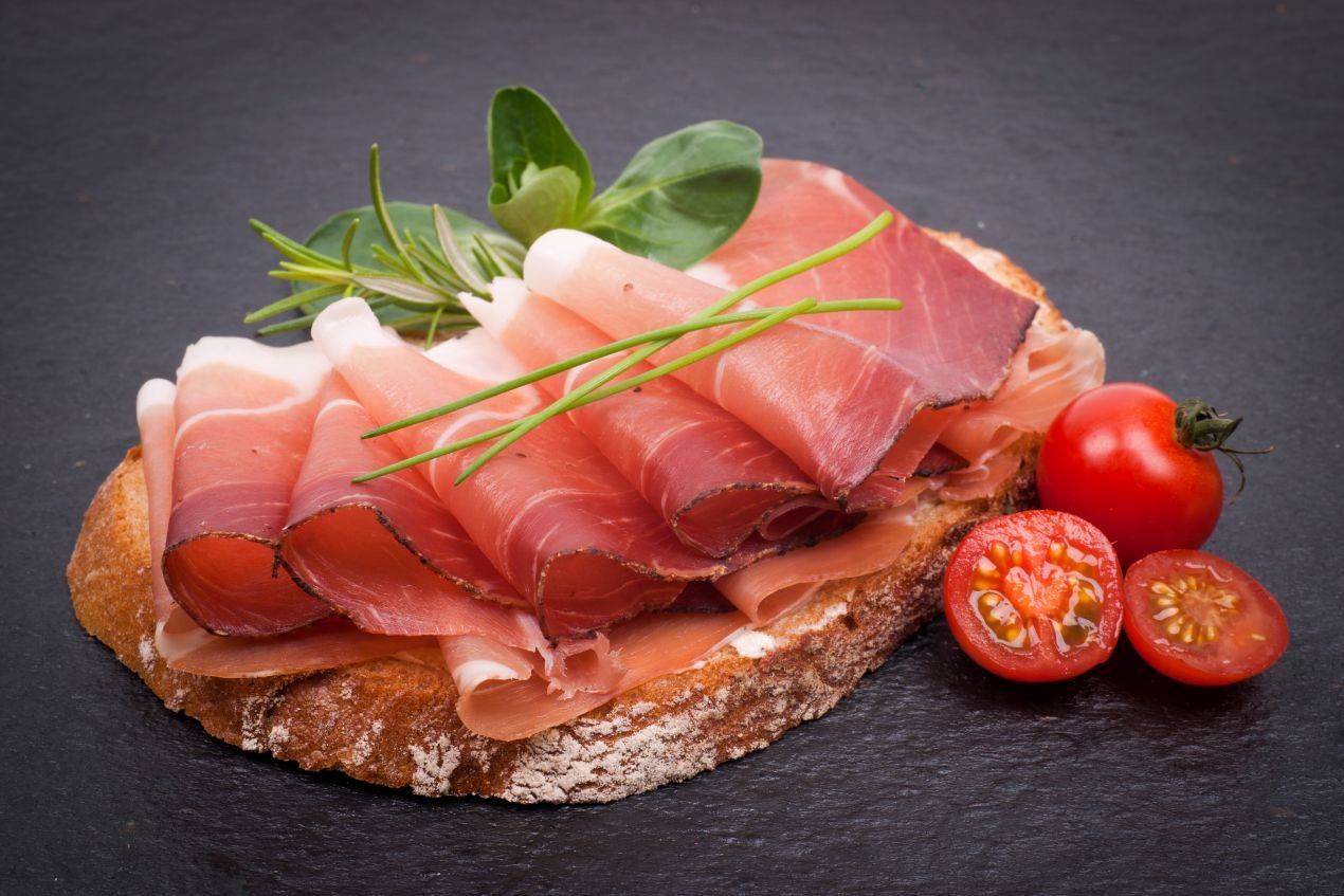 Hausgemachtes Brot mit fein aufgerolltem Schinkenspeck, Cocktailtomaten und frischen Kräutern – servierfertig dekoriert