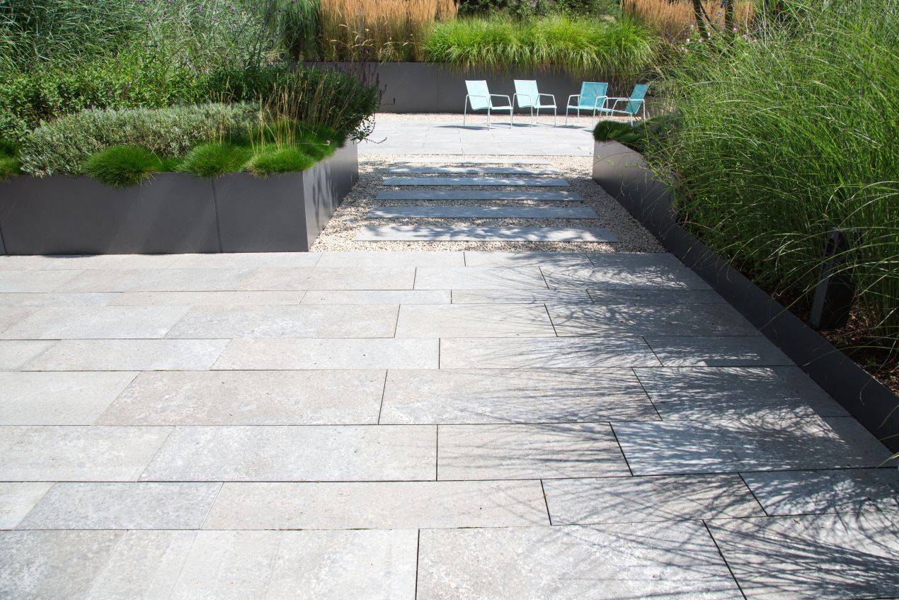 Helle Natursteinplatten auf einer modernen Terrasse mit Pflanzbeeten und Gartenstühlen, kombiniert mit Ziergräsern und klarer Linienführung